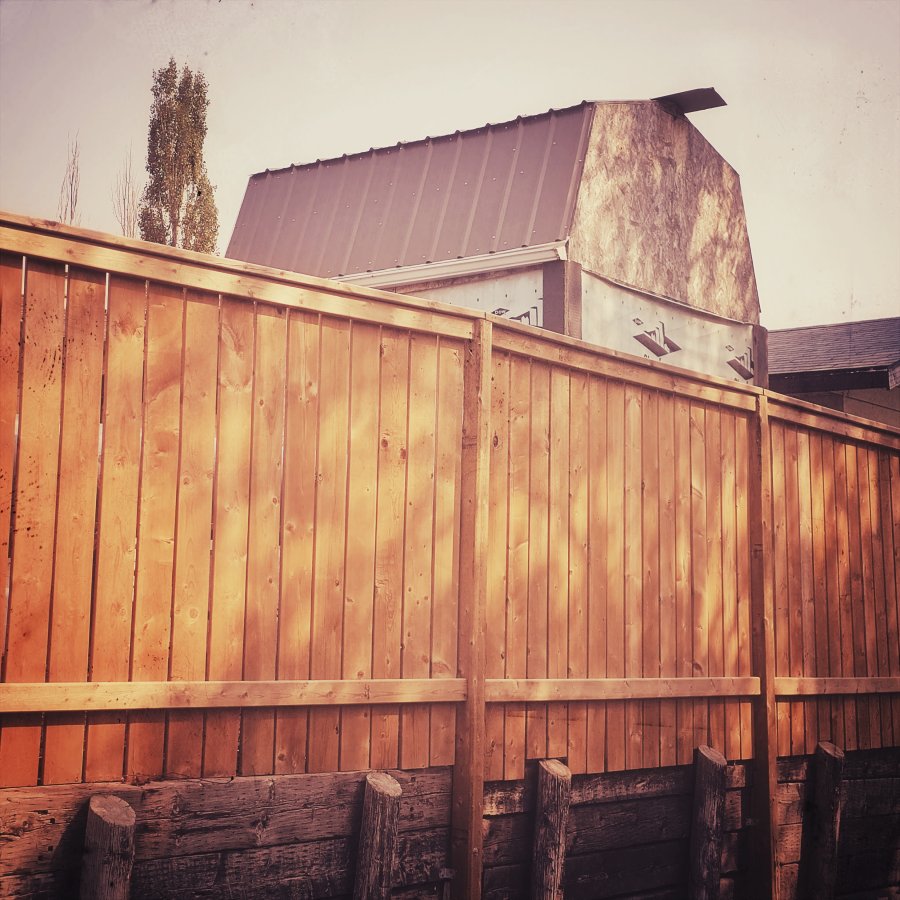 A light brown wooden fence stands on a timber retaining wall, with the roof of an unfinished barn and a tall tree visible in the background under a pale, hazy sky.