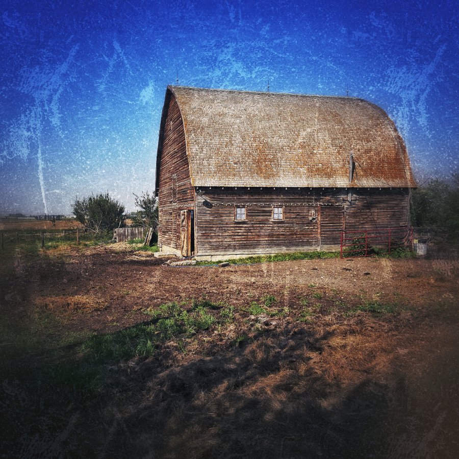 A rustic wooden barn with a high shingled roof stands in a dry dirt yard against a vivid blue sky, framed by a distressed vintage border.