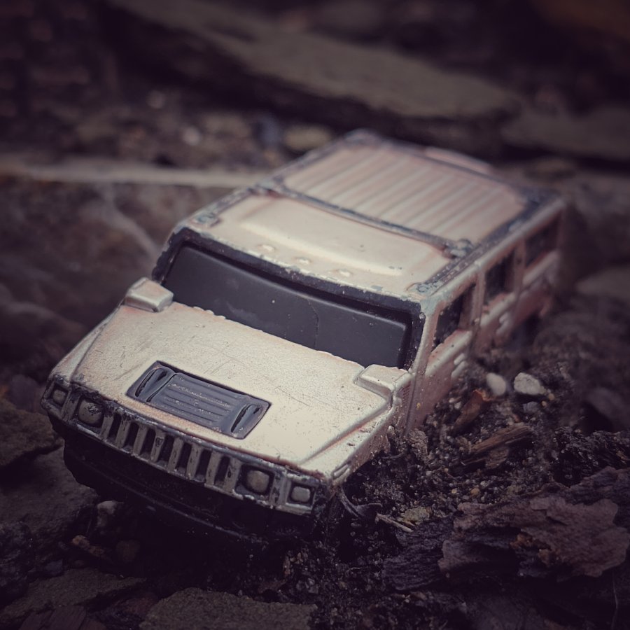 A miniature silver toy SUV is partially buried in dark, damp soil and wood bark.