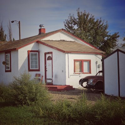 A small white stucco house with red trim has a rusted vintage car chassis parked in the dirt driveway next to a white shed.